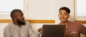 Two individuals are sitting at a table with a laptop, discussing something in a brightly lit room.